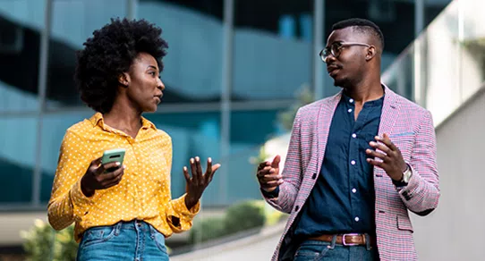 Two people are engaged in conversation outdoors. The woman, holding a phone, gestures expressively while the man, wearing glasses and a patterned blazer, listens attentively. A modern building is in the background.