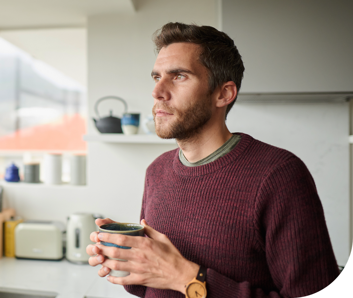 A man holding a mug gazes thoughtfully out a kitchen window. He wears a maroon sweater, surrounded by modern kitchen appliances and a warm, inviting atmosphere.