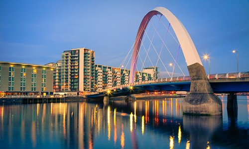 A modern arch bridge spans a reflective river, connecting urban structures on either side, with buildings illuminated under a twilight sky, creating a serene cityscape.