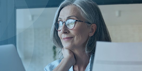A woman with grey hair and glasses gazes thoughtfully at a laptop while resting her hand on her neck, surrounded by a bright, modern workspace with soft natural light.