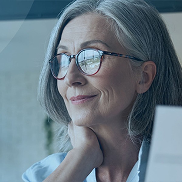 A woman with grey hair and glasses gazes thoughtfully at a laptop while resting her hand on her neck, surrounded by a bright, modern workspace with soft natural light.
