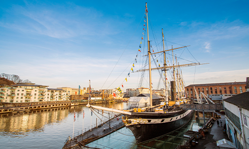 A tall sailing ship is docked at a marina, with colorful flags flying and reflections on the water. Modern buildings line the waterfront under a clear blue sky.