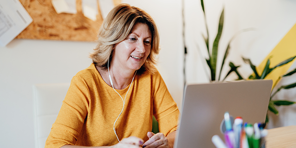 A woman in an orange sweater smiles while using a laptop. She wears earphones and sits at a desk cluttered with colorful pens, surrounded by plants and a clipboard display.