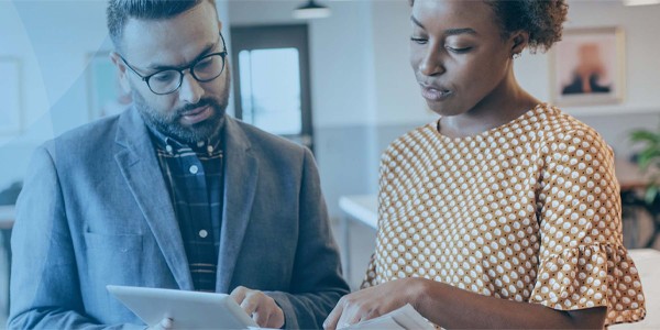A man and a woman are engaged in discussion; he is holding a tablet while she points at a document in her hand. The setting is a modern office with bright lighting.
