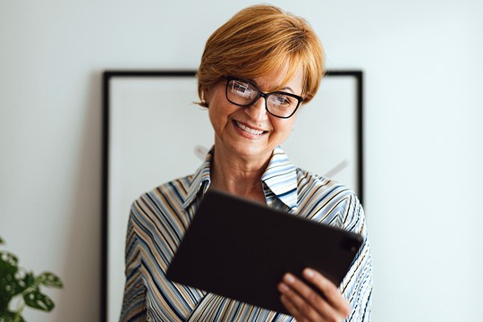 A woman with short, light brown hair smiles while holding a tablet. She is wearing a striped shirt and appears to be in a bright, minimalistic room, with a plant and framed artwork nearby.