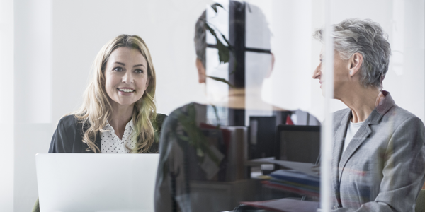 A woman with long hair smiles while seated at a desk with a laptop. Another woman in a gray blazer listens, surrounded by a bright office with plants and visible file storage.