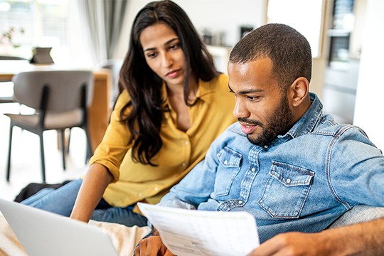A man and a woman sit closely together on a couch, reviewing documents while looking at a laptop. The room features modern furnishings and natural light, creating a relaxed atmosphere.