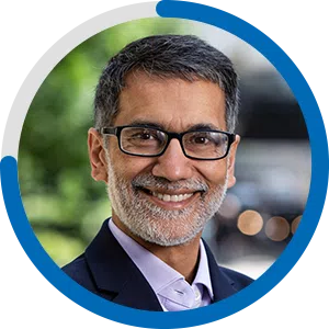 A man with glasses and a trimmed beard smiles warmly, surrounded by a soft-focus green background, indicating an outdoor setting with blurred lights suggesting a social atmosphere.