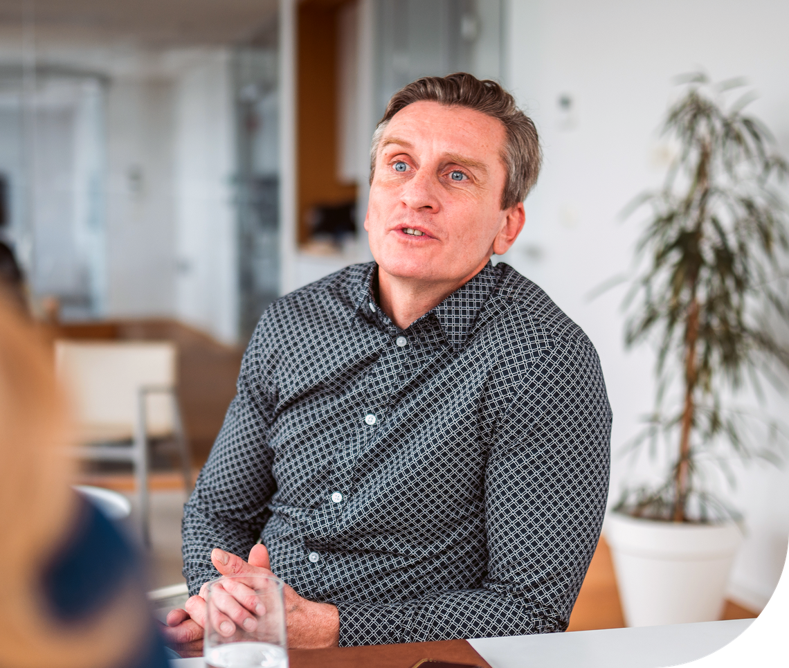 A man in a patterned shirt is speaking engagingly at a conference table in a modern office setting, with a plant visible in the background and a glass of water in front of him.