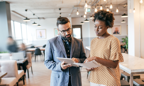A man and woman are discussing project materials while looking at a tablet and papers. They are in a contemporary café with tables and large windows, creating a bright atmosphere.