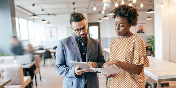 A man and woman are discussing project materials while looking at a tablet and papers. They are in a contemporary café with tables and large windows, creating a bright atmosphere.