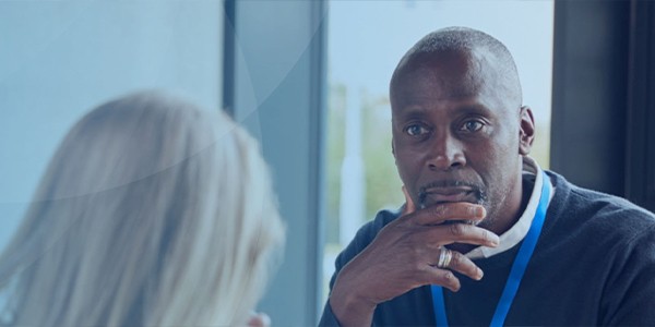 A man sits thoughtfully with his hand on his chin, engaging in conversation with a woman whose back is turned. The setting appears modern, with natural light filtering through large windows.
