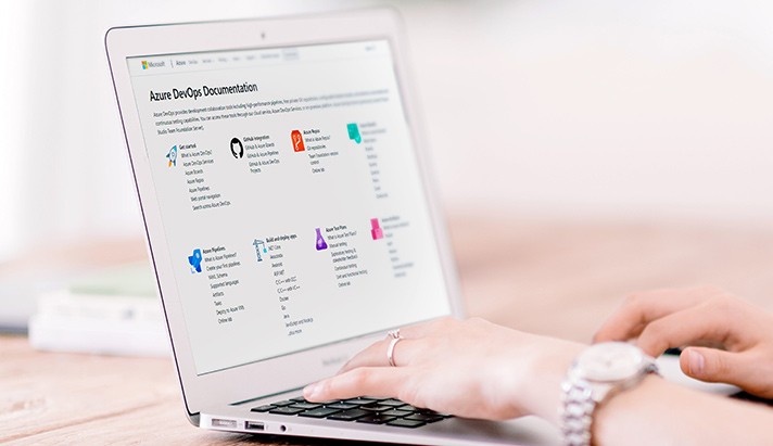 A person is using a laptop, browsing the Azure DevOps documentation website. The environment is a workspace with a wooden surface and a partially visible notebook in the background.