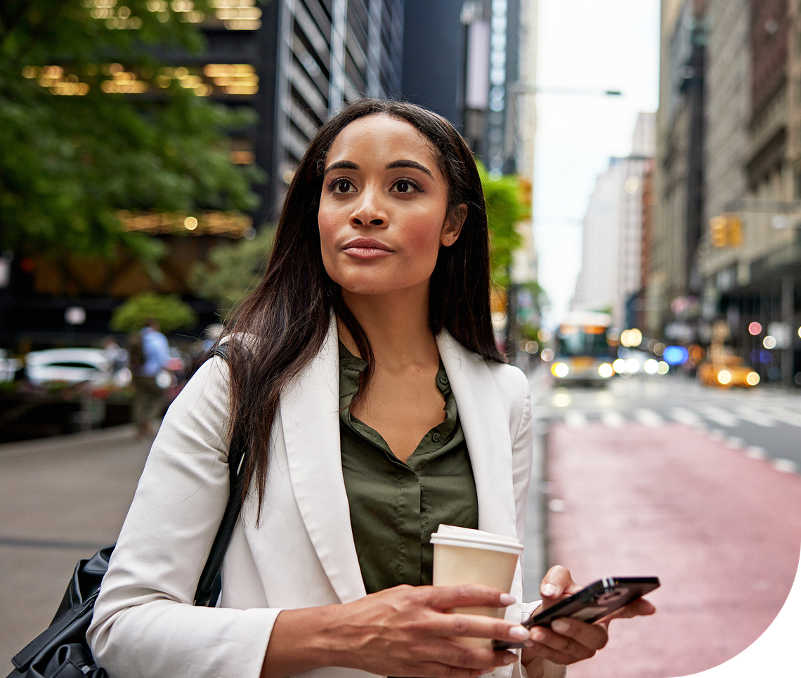A woman holding a coffee cup and a smartphone stands on a city street, appearing thoughtful, amidst tall buildings and bustling urban activity.