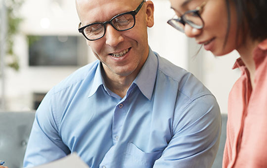 A man and a woman, both wearing glasses, are engaged in a discussion while examining documents. They are seated on a gray couch in a modern, brightly lit workspace with greenery in the background.