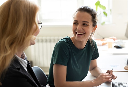 Two women are engaged in conversation, smiling warmly at each other. They're sitting in a bright office with a laptop and indoor plants in the background.