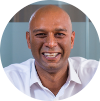 A bald man in a white shirt smiles broadly, sitting against a blurred, modern office background with glass panels, conveying a friendly and approachable demeanor.