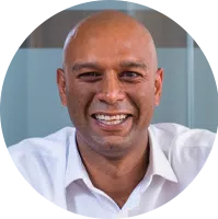 A bald man in a white shirt smiles broadly, sitting against a blurred, modern office background with glass panels, conveying a friendly and approachable demeanor.