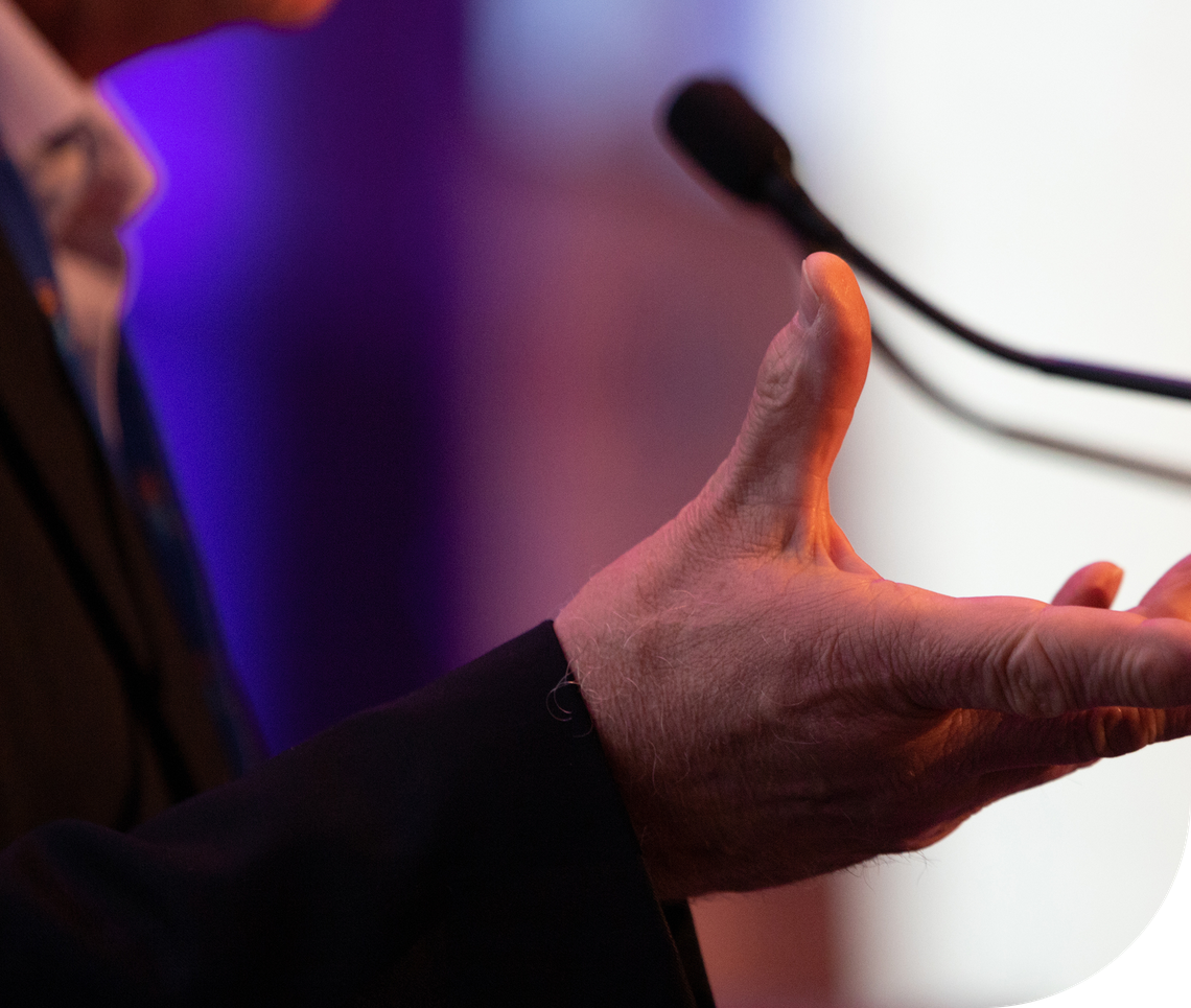 A hand gestures expressively while speaking into a microphone. The background features blurred purple and soft light tones, suggesting a formal presentation setting.