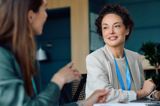 Two women sit across from each other in a modern office setting. One woman, smiling, listens attentively while the other gestures, engaged in conversation. Greenery is visible in the background.