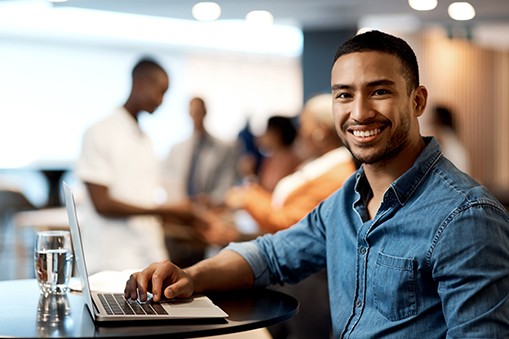 A young man sits at a table with a laptop, smiling at the camera. In the background, groups of people are engaged in conversation in a modern, well-lit environment. A glass of water is nearby.