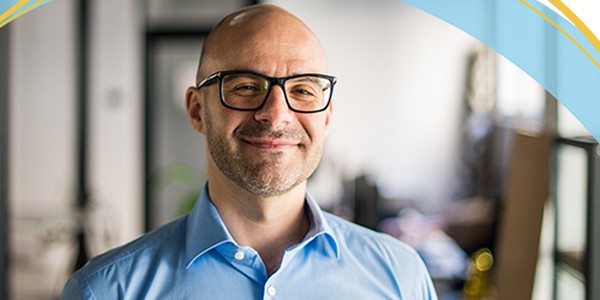 A smiling bald man wears glasses and a blue shirt, standing confidently in a bright, modern office with blurred furniture and plants in the background.