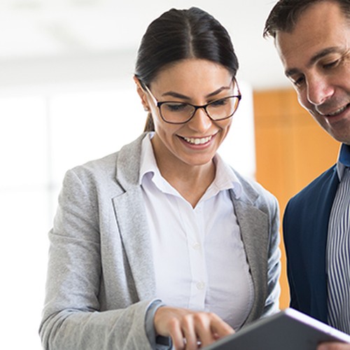 A woman in glasses and a gray blazer points at a tablet while smiling, engaging with a man in a blue suit. They are in a brightly lit office environment.
