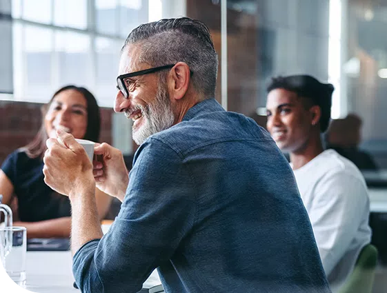 A smiling man holds a coffee cup while seated at a table with others. They are engaged in conversation in a bright, modern workspace with large windows and a relaxed atmosphere.