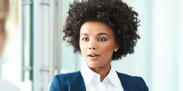 A young woman with curly hair looks thoughtfully at another person during a conversation in a modern, well-lit office environment, suggesting a professional discussion or interview setting.