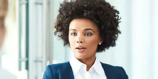 A young woman with curly hair looks thoughtfully at another person during a conversation in a modern, well-lit office environment, suggesting a professional discussion or interview setting.