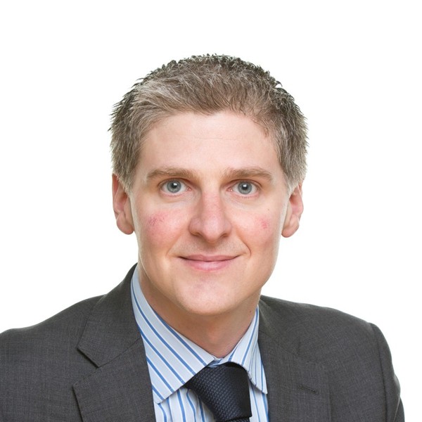 A man with short, light-colored hair smiles confidently. He wears a gray suit and a striped shirt with a tie, set against a plain white background.