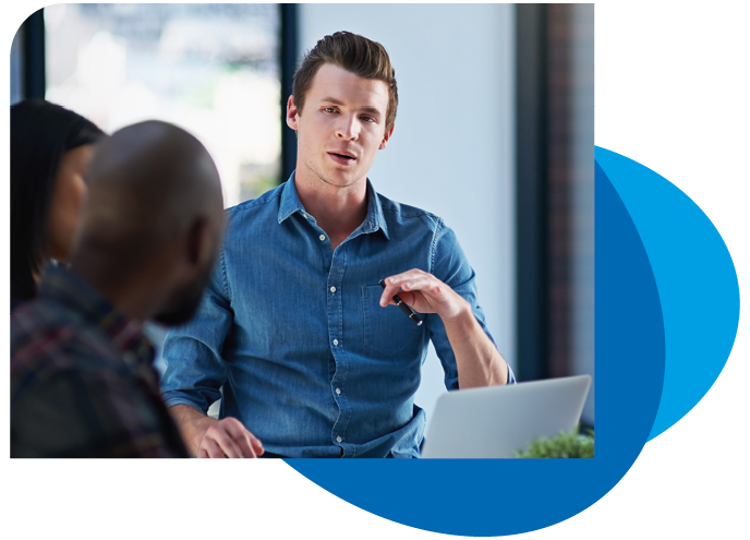 A young man in a blue shirt is speaking animatedly while gesturing, engaging with two people in a modern office setting featuring large windows and a laptop on the table.