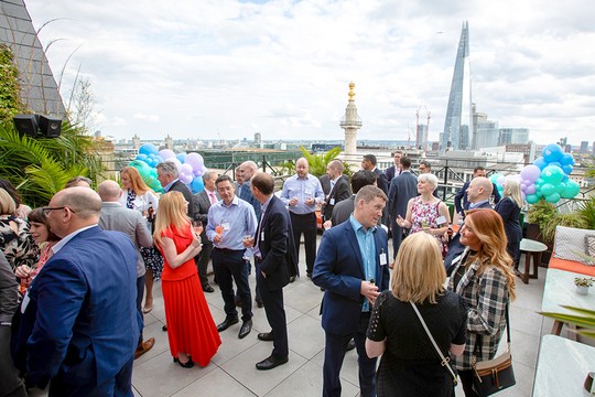A group of people mingles on a rooftop terrace, holding drinks and chatting. The scene features colorful balloons and a skyline backdrop with prominent buildings, including The Shard.