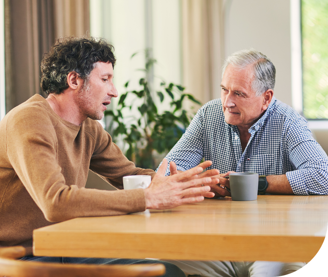 Two men engage in a conversation at a wooden table, one animatedly gesturing while the other listens attentively, in a bright, contemporary room with a plant in the background.