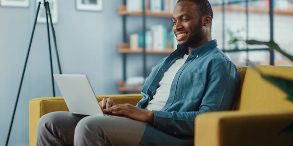 A man sits on a yellow couch, smiling while using a laptop. Behind him is a bookshelf filled with books, and a floor lamp stands nearby in a cozy living space.