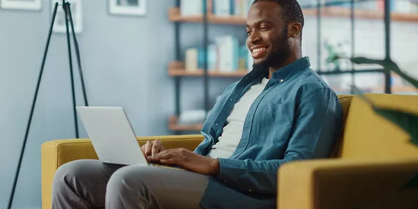 A man sits on a yellow couch, smiling while using a laptop. Behind him is a bookshelf filled with books, and a floor lamp stands nearby in a cozy living space.