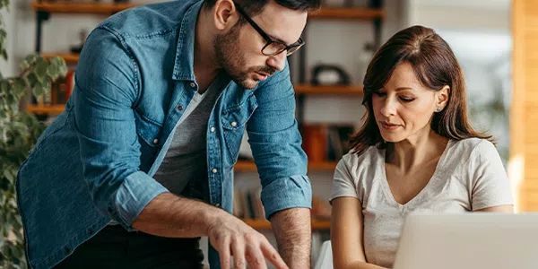 A man in a denim jacket gestures towards a laptop while discussing with a woman in a light top. They are seated at a table in a bright, modern room with bookshelves.