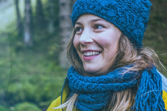 A smiling woman wearing a blue knitted hat and scarf stands outdoors, surrounded by lush greenery. She appears happy and engaged in her environment, suggesting a serene moment in nature.
