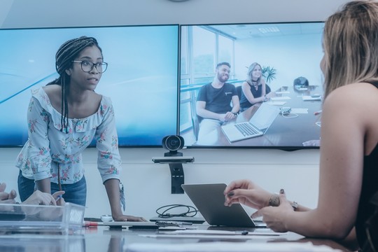 A group of diverse individuals engages in a meeting around a table, with one person presenting to others. A large screen displays remote participants, enhancing collaboration in a modern office environment.