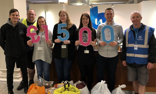 A group of six people stands in an office holding large numbers that total £1,500, surrounded by bags of donations on the floor. The setting conveys a charitable event.