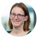 A woman with glasses and shoulder-length brown hair smiles warmly. She is wearing a green shirt, with a modern office environment visible in the blurred background.