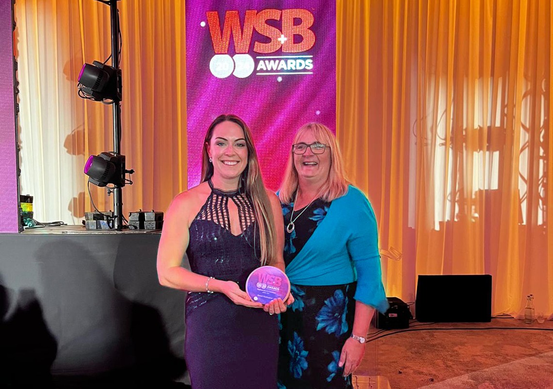 A woman in elegant attire holds an award, smiling alongside another woman at a brightly lit event featuring a backdrop displaying "WSB Awards 2023." The atmosphere appears celebratory.
