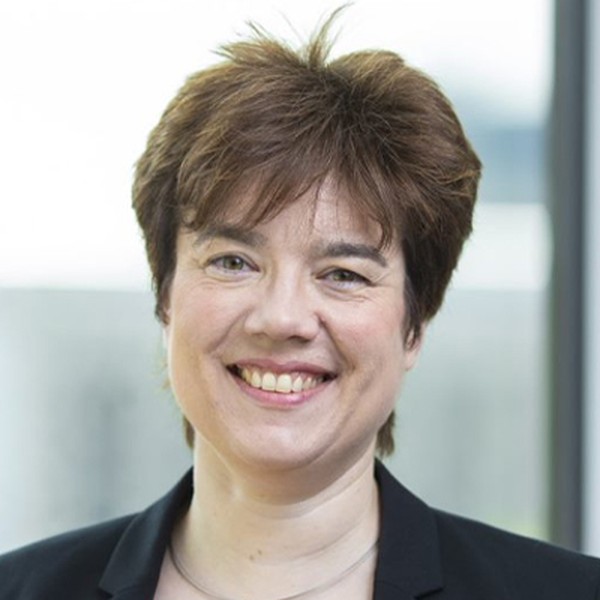 A person with short brown hair and a friendly smile is facing forward, wearing a black blazer against a bright, blurred background suggesting an indoor setting with natural light.