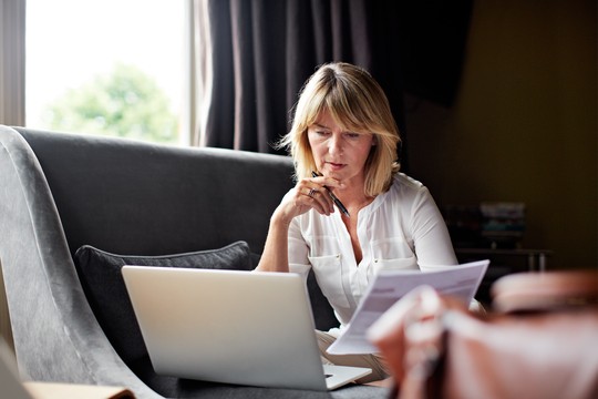 A woman sits on a gray couch, focused on her laptop while holding a pen and reviewing documents. Natural light filters in through a window, creating a cozy atmosphere.