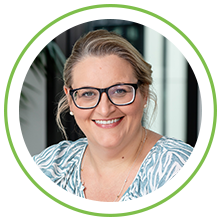 A smiling woman with glasses and wavy hair is posed in a circular frame. She wears a blue and white patterned top, seated in a well-lit indoor environment with greenery.