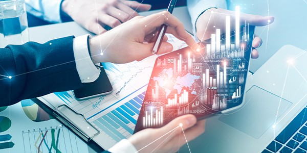 A tablet displaying digital graphs and data is held by two hands. One hand is pointing at the screen while the other hand writes with a pen, surrounded by financial documents and a laptop.