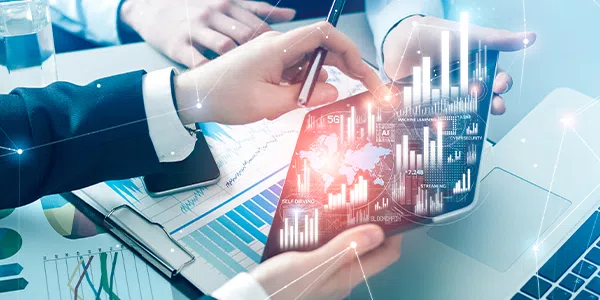 A tablet displaying digital graphs and data is held by two hands. One hand is pointing at the screen while the other hand writes with a pen, surrounded by financial documents and a laptop.