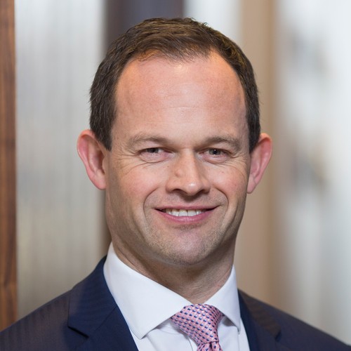 A man in a suit smiles confidently at the camera, with a blurred indoor background featuring wood tones and soft lighting, suggesting a professional or business setting.