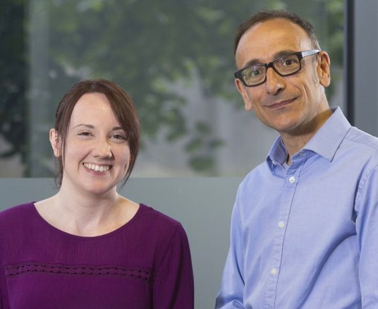 Two individuals are smiling while posing for a photo in a well-lit indoor setting, with greenery visible through the window behind them. One wears a purple top, and the other a blue shirt.