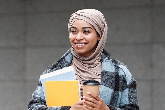 A woman wearing a beige hijab and a checkered coat holds notebooks and a coffee cup, smiling while standing against a muted, textured wall background.